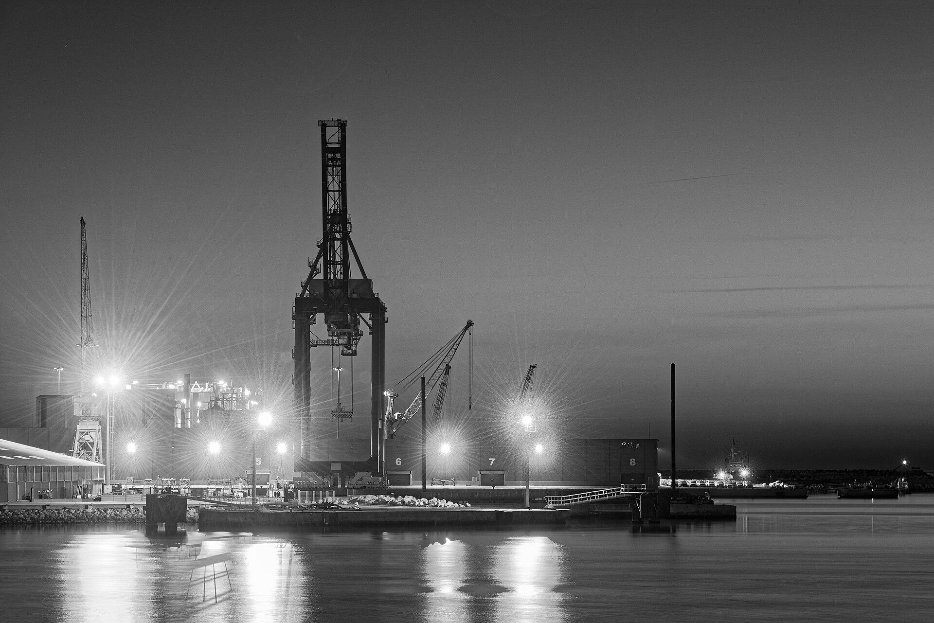 Harbour cranes and quay lights reflected in still water after dark.